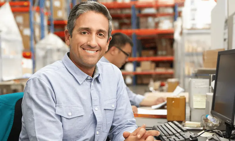 A middle-aged person in a light blue button-up shirt sits in front of a computer in a warehouse setting. The person is smiling and looking at the camera.